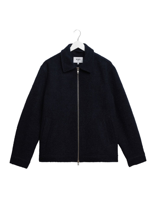A dark navy blue zip-up jacket with a textured bouclé-like fabric, featuring a collar and a silver-tone front zipper, hanging on a white hanger against a plain white background.